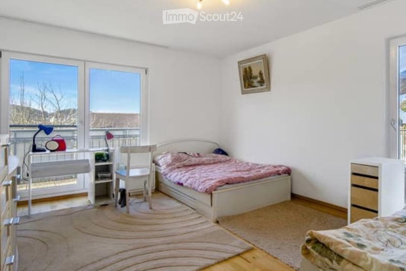 Cozy bedroom with a double bed, desk, chair, and large windows offering a scenic view. The room features a round rug, a nightstand, and a mirror on the wall.