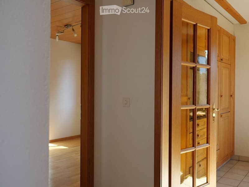 A well-lit hallway with wooden flooring, light-colored walls, and a wooden door with glass panels leading to another room.
