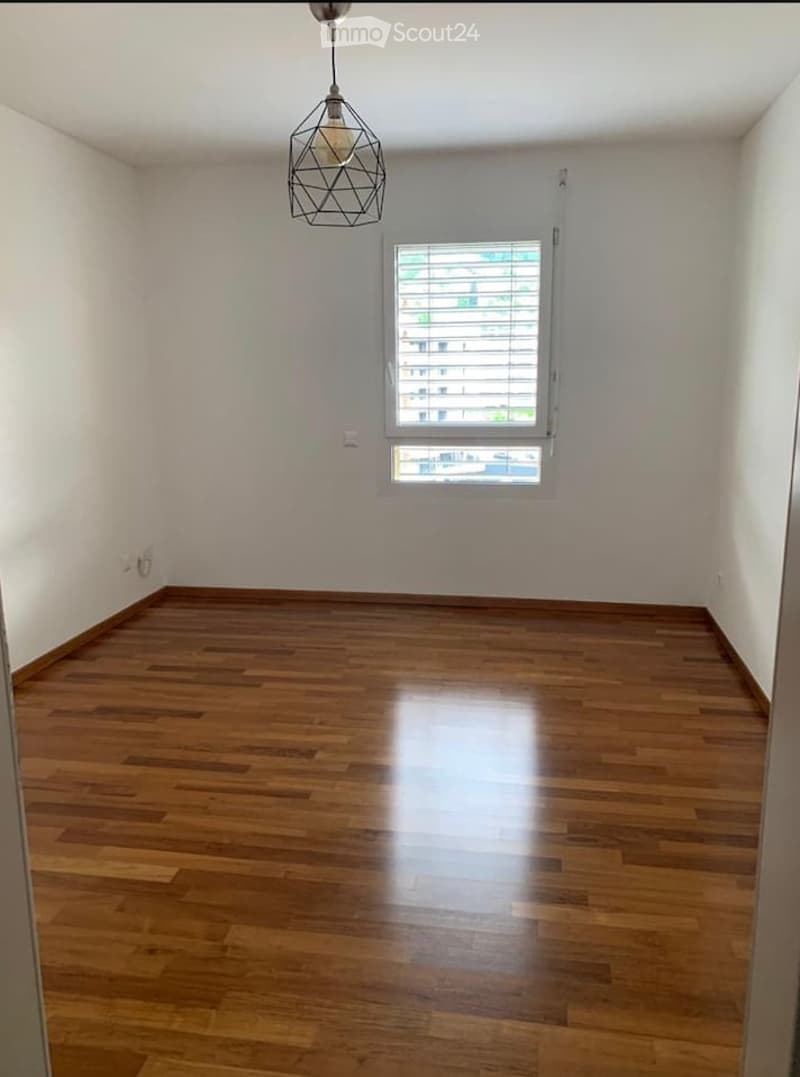 Empty room with wooden flooring, white walls, and a single window with blinds. A modern pendant light hangs from the ceiling.