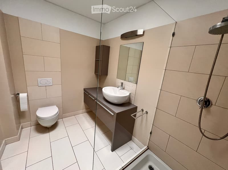 Modern bathroom with beige tiles, a toilet, a sink with a mirror, a shower, and a cabinet.