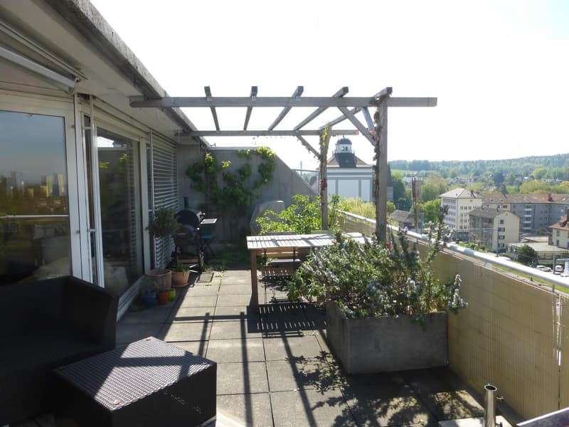 Balcony with a wooden pergola, sofa, table, bench, potted plants, and a grill, overlooking a cityscape with buildings and greenery.
