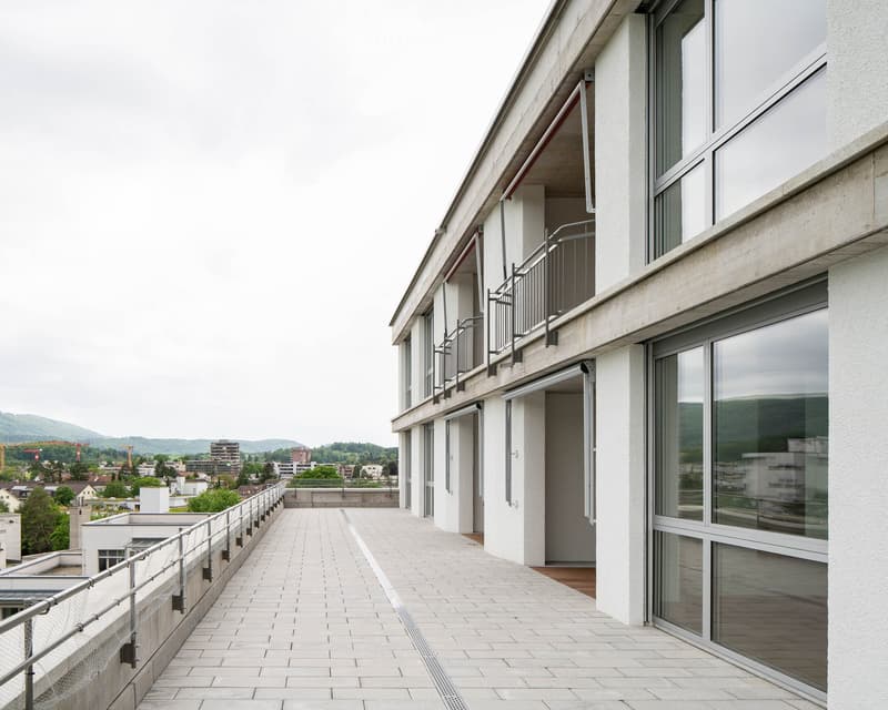 Modern balcony area with white tiled flooring, metal railings, and large glass windows overlooking a cityscape with distant mountains.