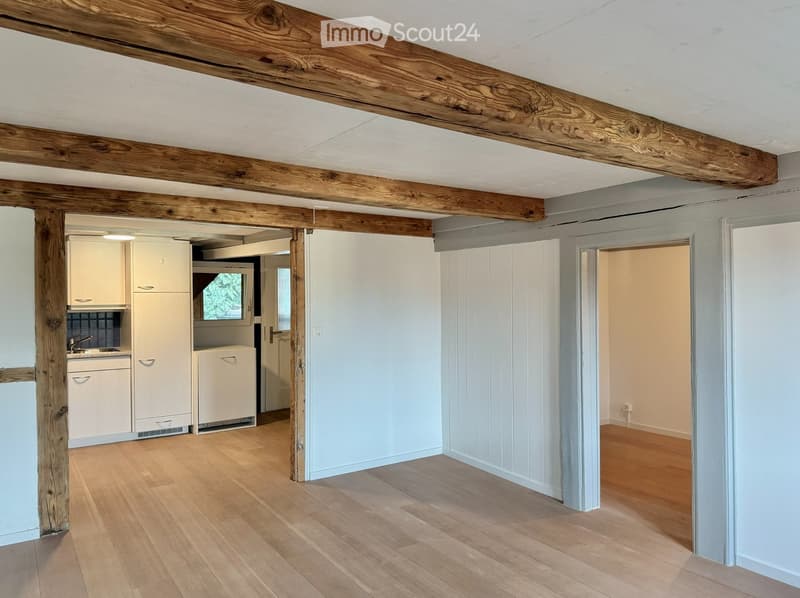 Bright, open room with wooden beams and light wooden flooring. A small kitchen area is visible with white cabinets, a sink, and a refrigerator. The space is clean and well-lit.
