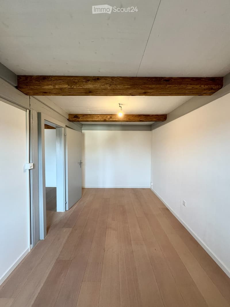 Empty room with wooden flooring, white walls, and exposed wooden beams on the ceiling. A single light fixture is visible, and there is a doorway leading to another room.