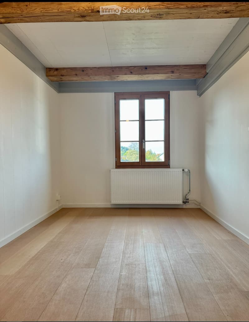 Empty room with light wooden flooring, white walls, a window with wooden frame, and a radiator below it.