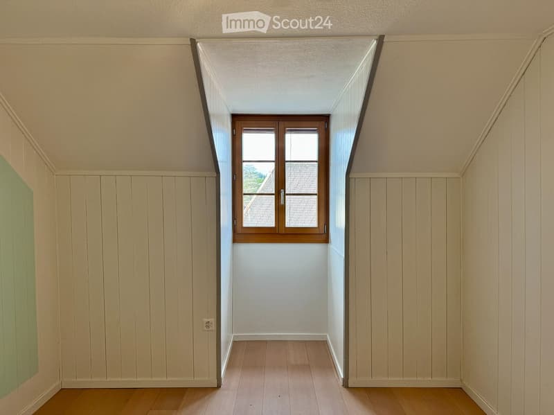 Empty attic space with wooden flooring, white walls, and a single window with wooden frame and glass panels.