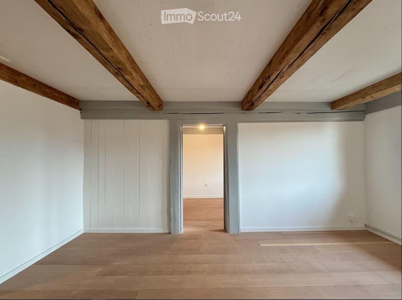 Empty room with wooden floor, white walls, and exposed wooden beams on the ceiling. A doorway leads to another room.