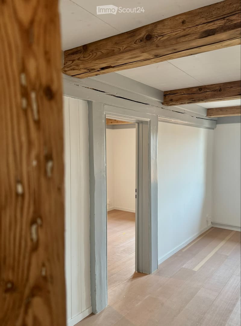 Empty hallway with light wooden flooring, white walls, and exposed wooden beams on the ceiling. A doorway leads to another room with similar flooring.