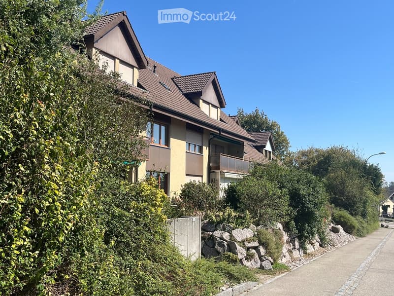 Two-story house with a brown tiled roof, large windows, and a balcony on the upper level, surrounded by greenery and a stone retaining wall.