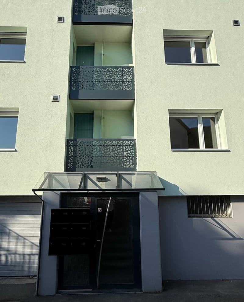 Modern multi-story building with light-colored facade, multiple balconies with decorative railings, and a glass-covered entrance area.