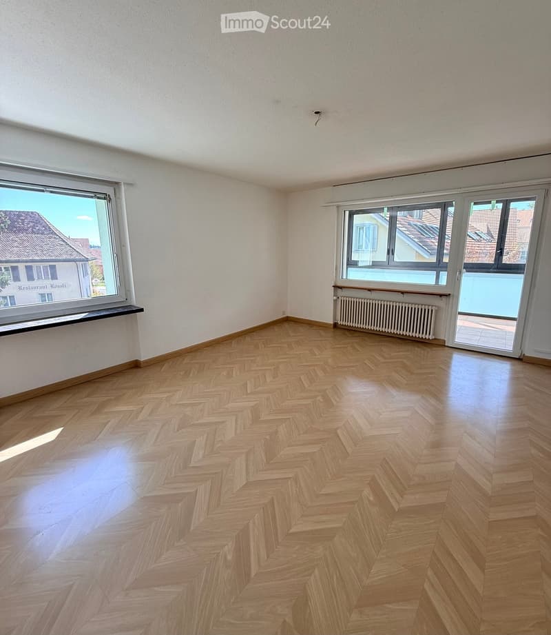 Empty room with herringbone wooden flooring, white walls, large windows, and a radiator. No furniture or equipment present.