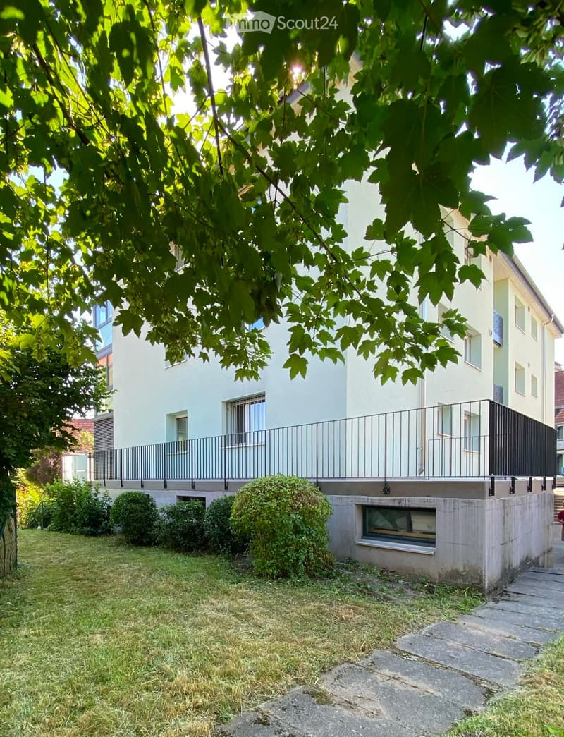 A well-maintained garden area with a modern two-story house in the background, featuring a balcony with metal railings, neatly trimmed bushes, and a stone pathway leading to the entrance.