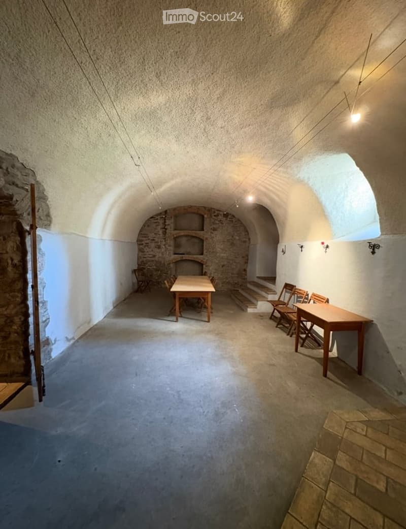 A rustic, arched room with stone walls and a concrete floor, featuring wooden tables and chairs arranged in the space.