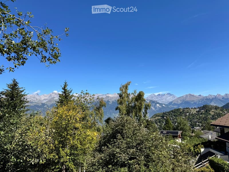 A scenic outdoor view featuring lush green trees in the foreground and a picturesque mountain range in the background under a clear blue sky.