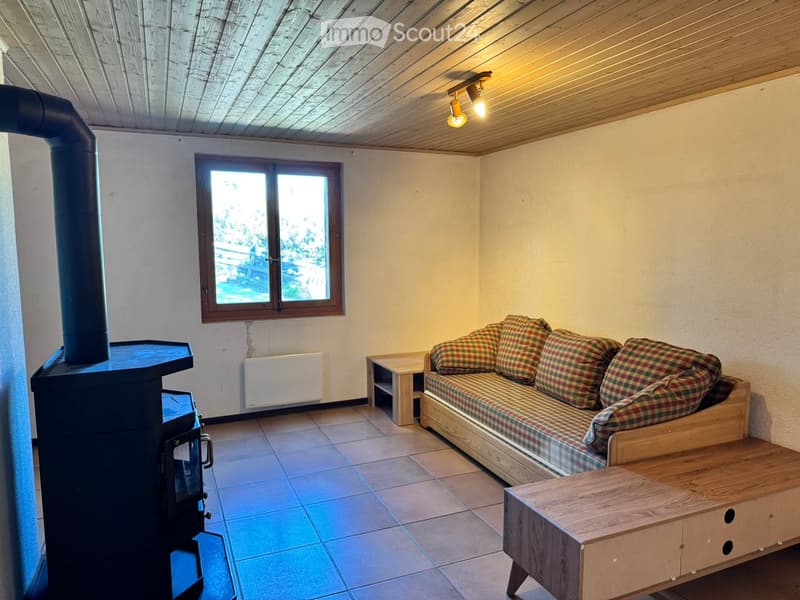 Cozy living room with a wooden stove, plaid-patterned sofa, coffee table, and wooden side table. The room features a large window and warm lighting.