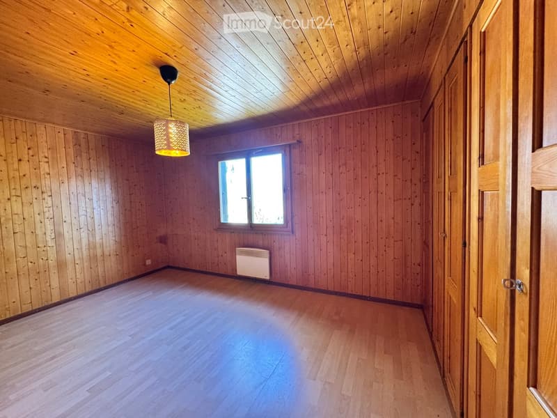 Empty room with wooden walls, ceiling, and floor, featuring a window, a radiator, and a hanging light fixture.