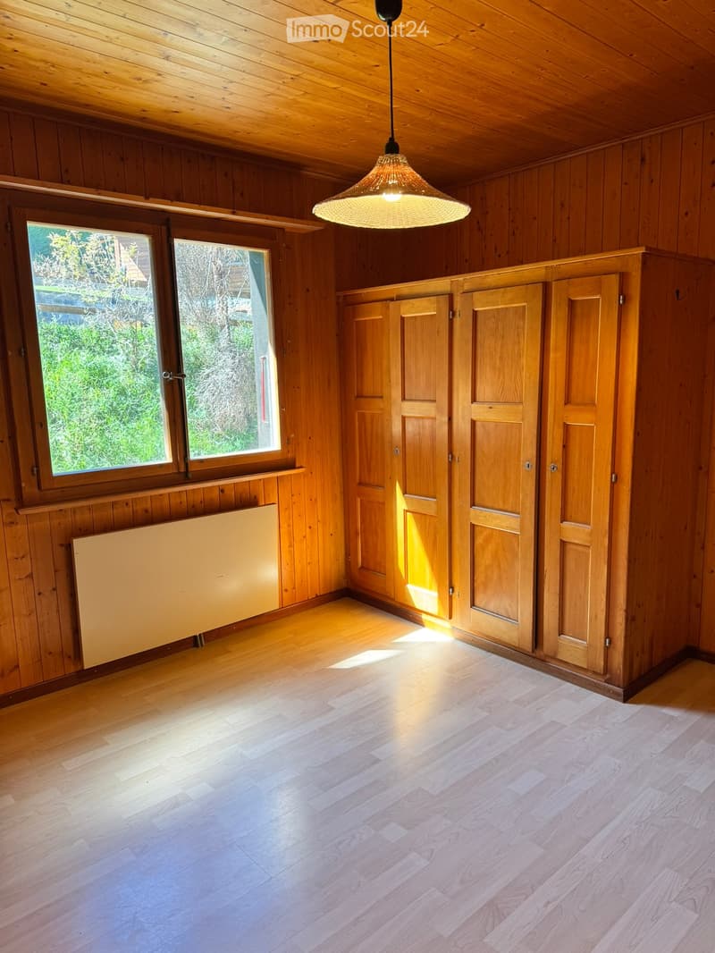 Empty room with wooden walls, wooden floor, large windows, built-in wooden cabinets, and a hanging light fixture.