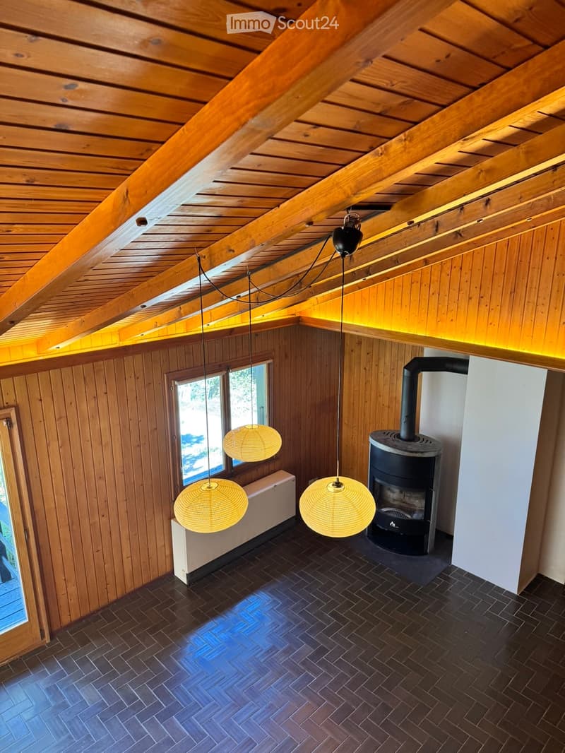 The room features exposed wooden beams and walls, a dark tiled floor, and a wood-burning stove with a chimney. Three woven pendant lights hang from the ceiling, and there is a window with a view of greenery outside.