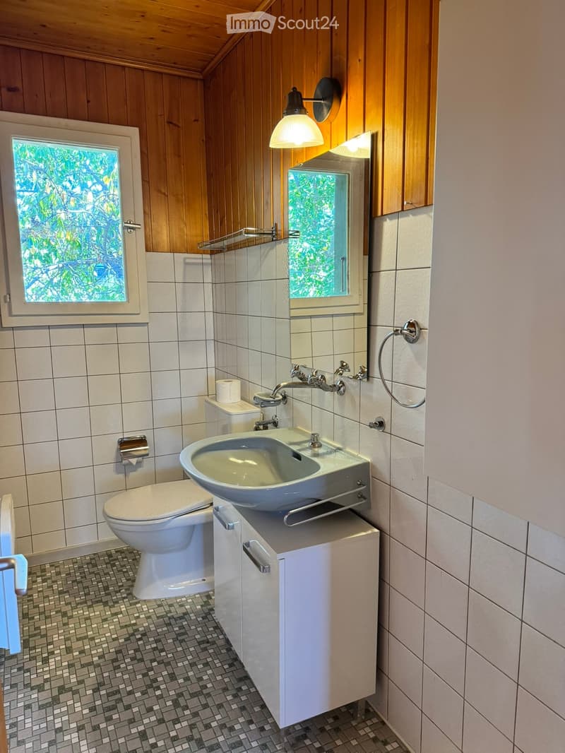 Modern bathroom with white tiles, wooden accents, a toilet, a sink with a modern faucet, a mirror, and a small cabinet. The room features a window with a view of greenery and a wall-mounted light fixture.