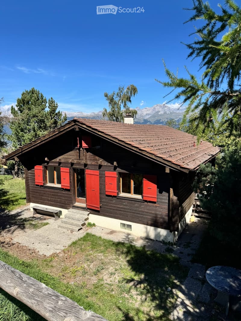 A wooden chalet with red shutters, situated in a mountainous area with trees and a clear blue sky.