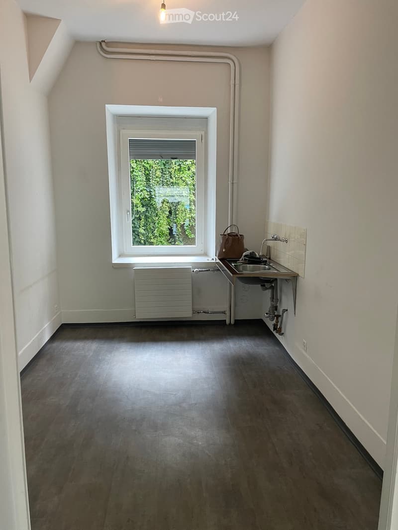 Small room with a window, sink, and a handbag placed on the floor. The room has a dark floor and white walls.
