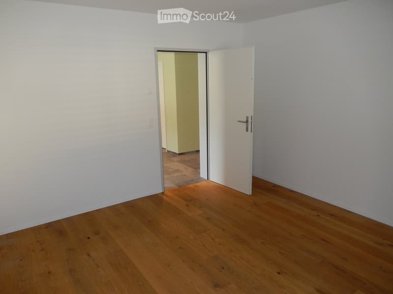 Empty room with wooden flooring and white walls, featuring an open doorway leading to another room.