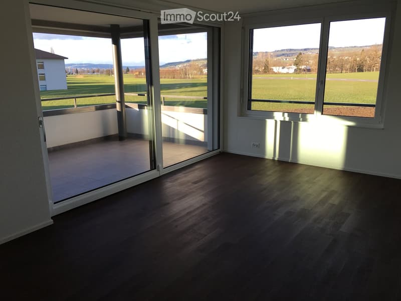 Empty room with wooden flooring, large windows, and a sliding glass door leading to a balcony with a scenic view of a green field and distant hills.