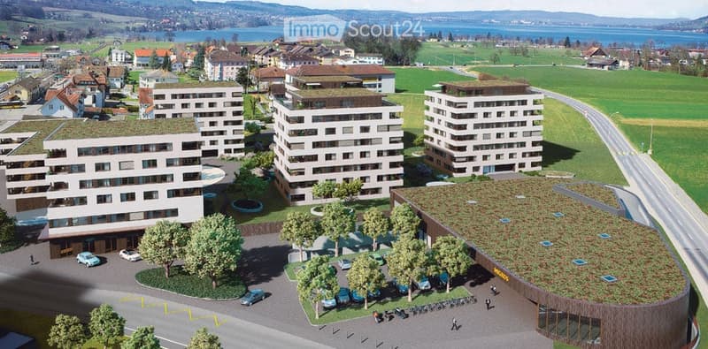 Aerial view of a modern residential complex with multiple apartment buildings, green spaces, parking area, and surrounding landscape with a lake and hills in the background.