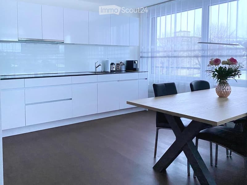 Modern kitchen with white cabinets, a sink, a coffee maker, and a dining area with a table and chairs. A vase with flowers is placed on the table.