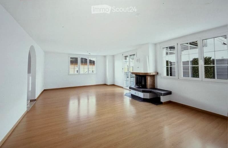 Empty room with wooden flooring, white walls, large windows, and a fireplace with a bench.