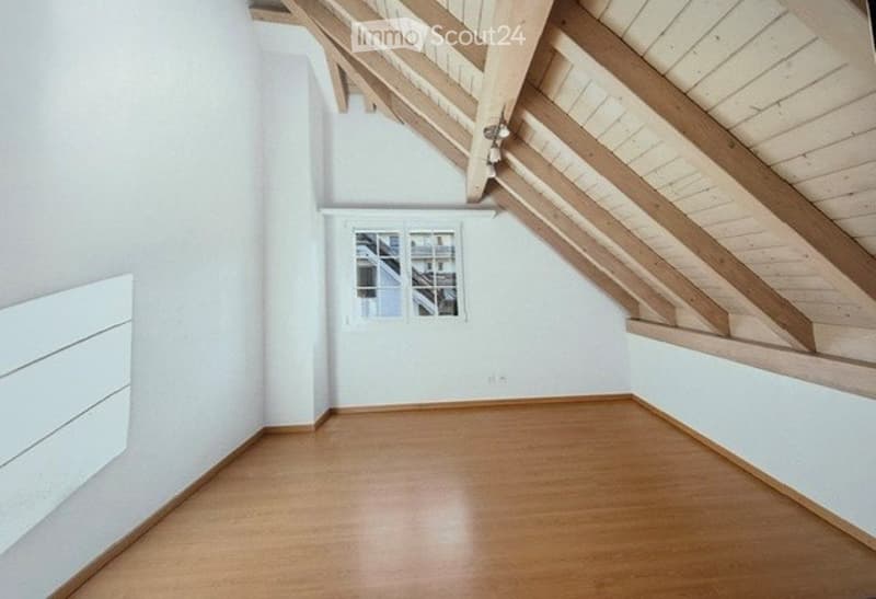 Empty attic room with wooden floor, white walls, exposed wooden beams, and a single window with white frame.