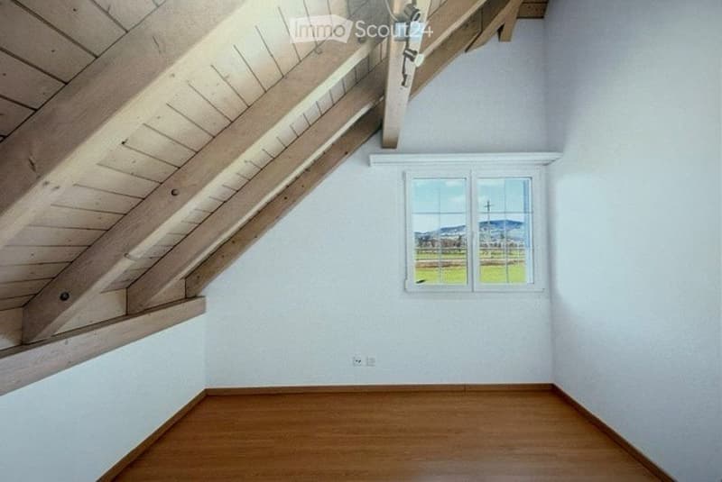 Empty room with wooden floor, white walls, exposed wooden beams on the ceiling, and a window offering a scenic view of a field and mountains.