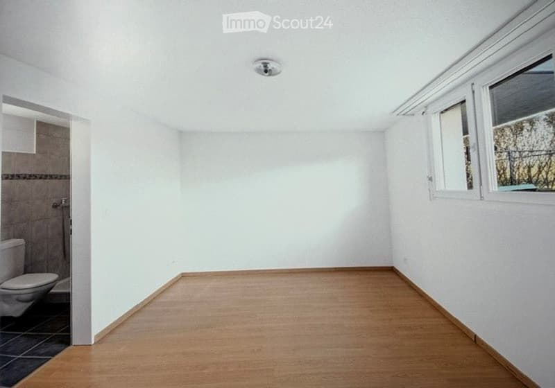 Empty room with white walls, wooden floor, and a small adjacent bathroom area visible through an open doorway.