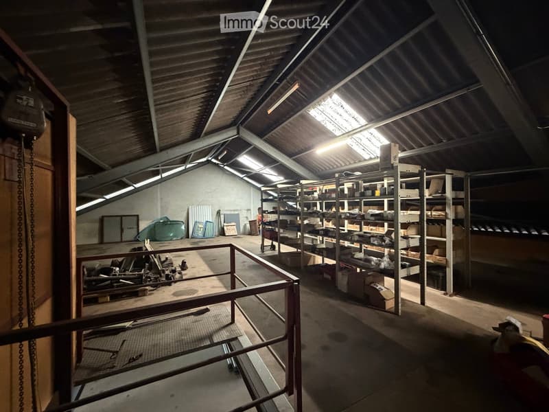 A spacious industrial-style storage area with metal shelving units, boxes, and tools scattered around. The space features a high, slanted metal roof with skylights and a metal railing on the upper level.