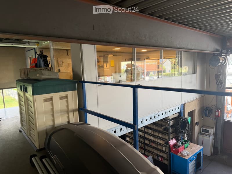 Workshop or garage area with a large storage unit, shelves, a workbench with tools, and a microwave. Visible through large windows is an outdoor area with a house and greenery.