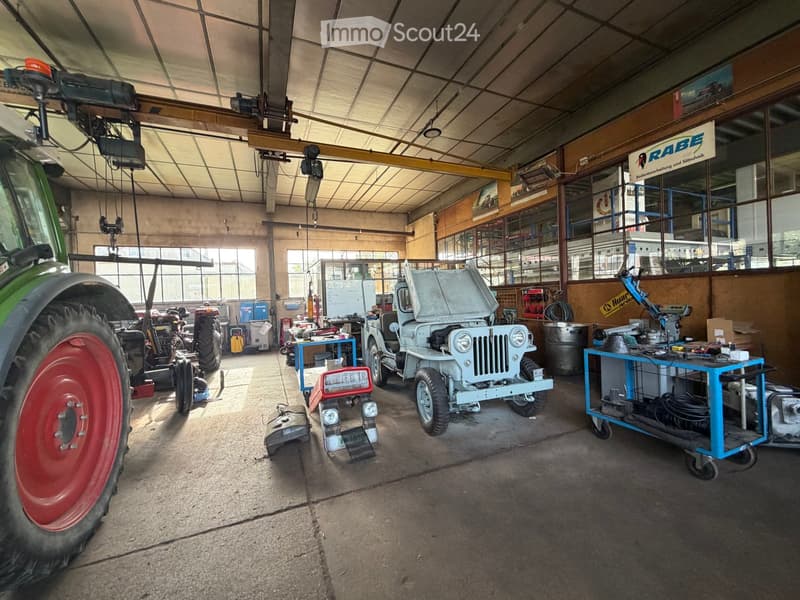 A spacious garage with a high ceiling, featuring a tractor, a small jeep, work tables, and various tools. The space is well-lit with natural light from large windows and has a crane system suspended from the ceiling.