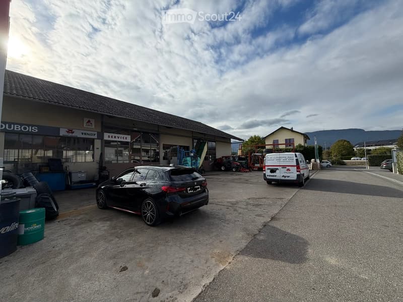 A commercial building with a sign reading "Dubois" and "Service" visible, with several vehicles including a black car, a white van, and agricultural machinery parked in front.