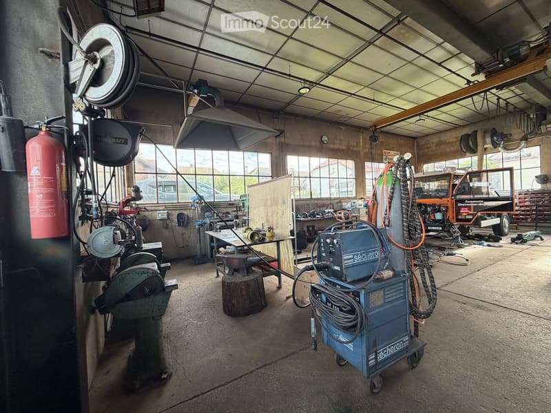 A spacious workshop with large windows, concrete floor, and various tools and equipment including a welding machine, grinder, workbench, and a truck in the background.