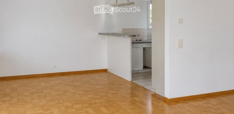 Empty room with wooden flooring, white walls, and a small kitchen area with cabinets and a countertop.