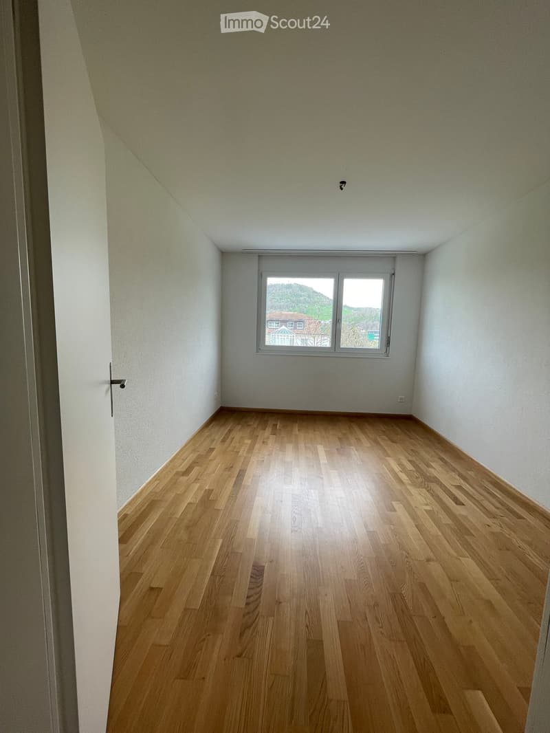 Empty room with light wooden flooring and white walls, featuring a large window with a view of greenery and a house outside.