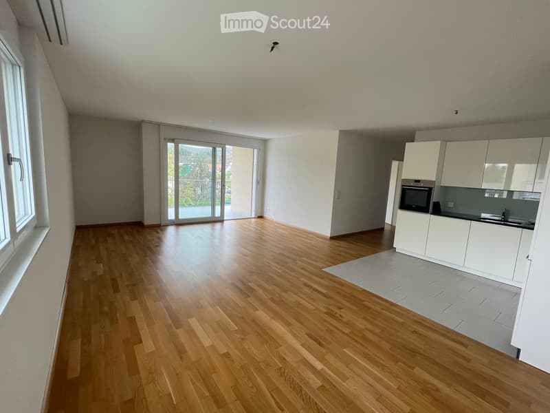 Empty room with wooden flooring, white walls, large windows, and a modern kitchen area with white cabinets and stainless steel appliances.