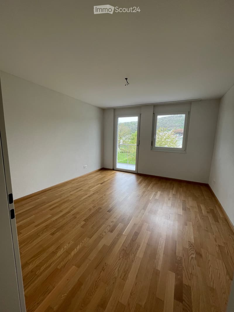 Empty room with wooden flooring, white walls, and large windows with a view of greenery outside.