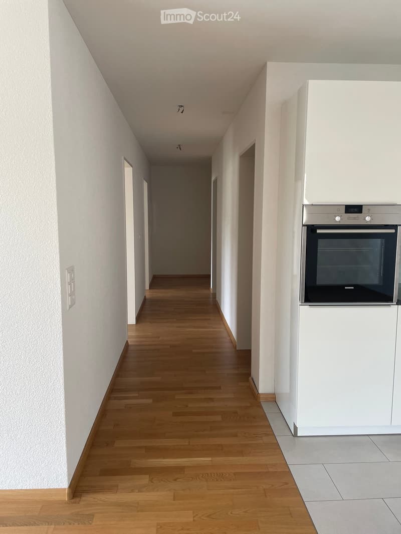 A narrow hallway with wooden flooring, white walls, and a modern oven in the corner. The hallway has several doors leading to other rooms.