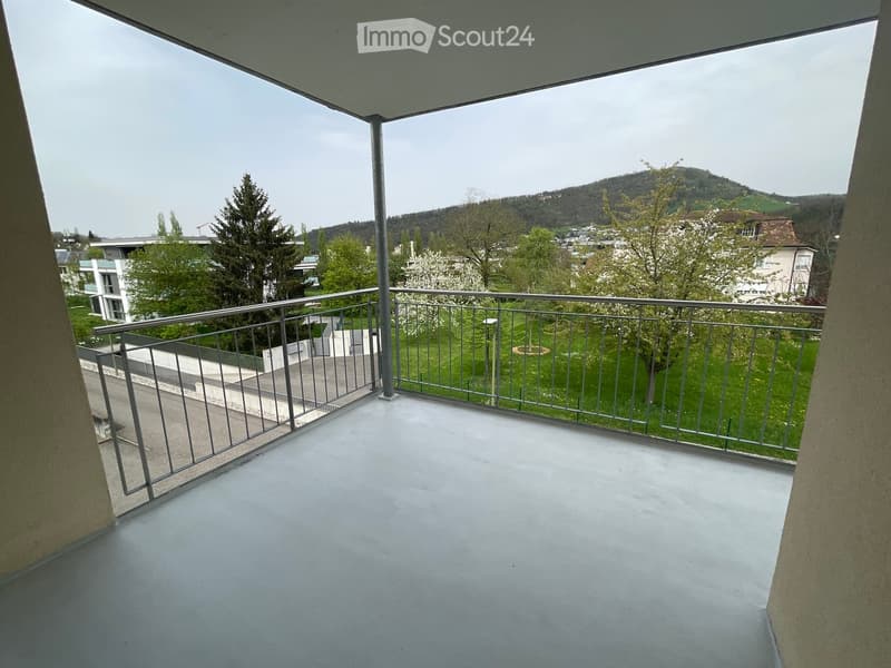 Balcony with metal railing, overlooking a green area with trees and a distant hill.