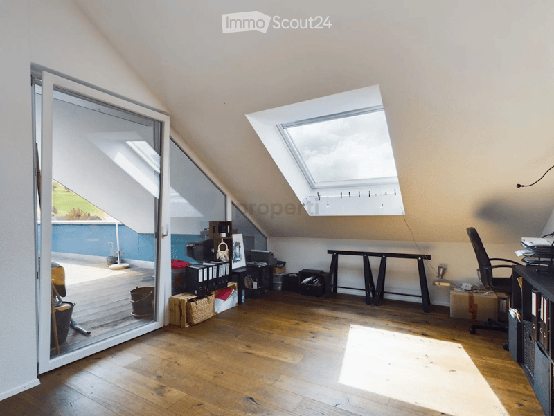 Bright attic room with wooden flooring, large skylight, and sliding glass door leading to a balcony. Contains a desk, chair, bookshelf, storage boxes, and a small table with a lamp.