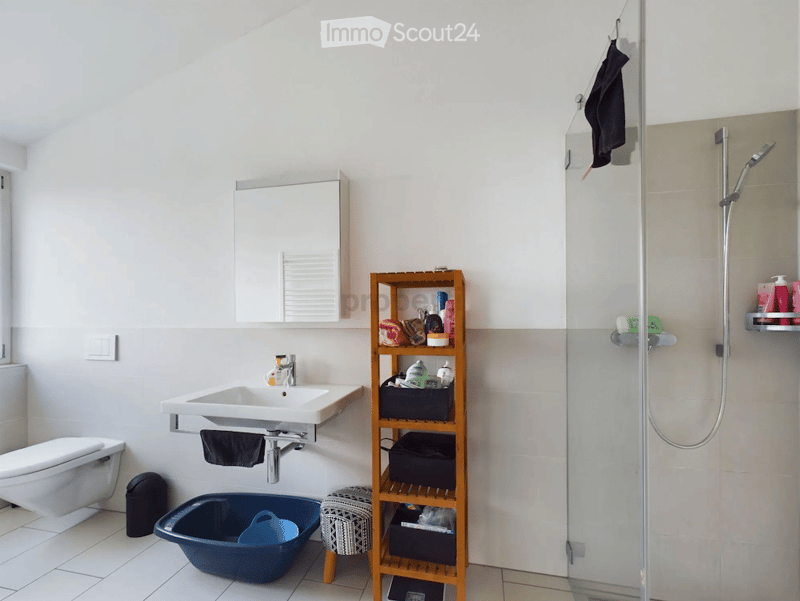 Modern bathroom with white tiled walls and floor, featuring a sink with mirror, toilet, shower with glass enclosure, wooden shelf with various items, and a small blue bathtub.