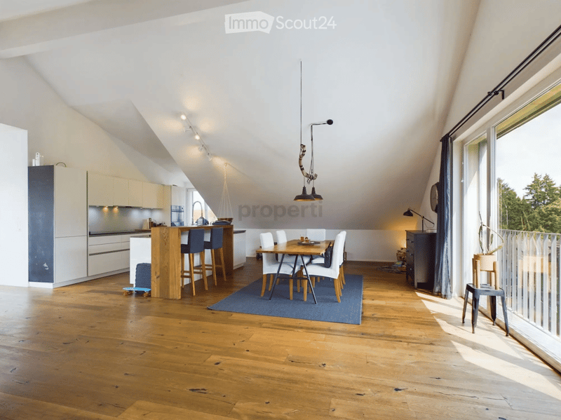 Open-plan kitchen and dining area with modern white cabinets, a central island, bar stools, a dining table with chairs, and a hanging pendant light. The space features wooden flooring and large windows letting in natural light.