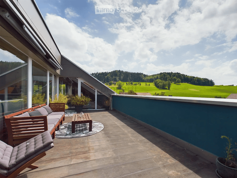 Balcony with wooden furniture, including a sofa, coffee table, and chair, overlooking a scenic countryside view.