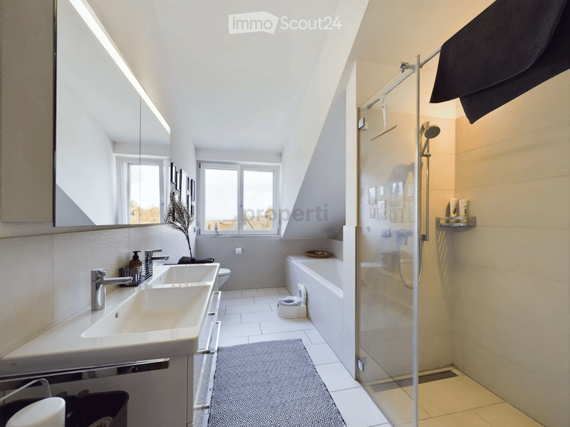 Modern bathroom with a large mirror, double sink, bathtub, and walk-in shower. Features include a towel rack, a small plant, and a window with a view.