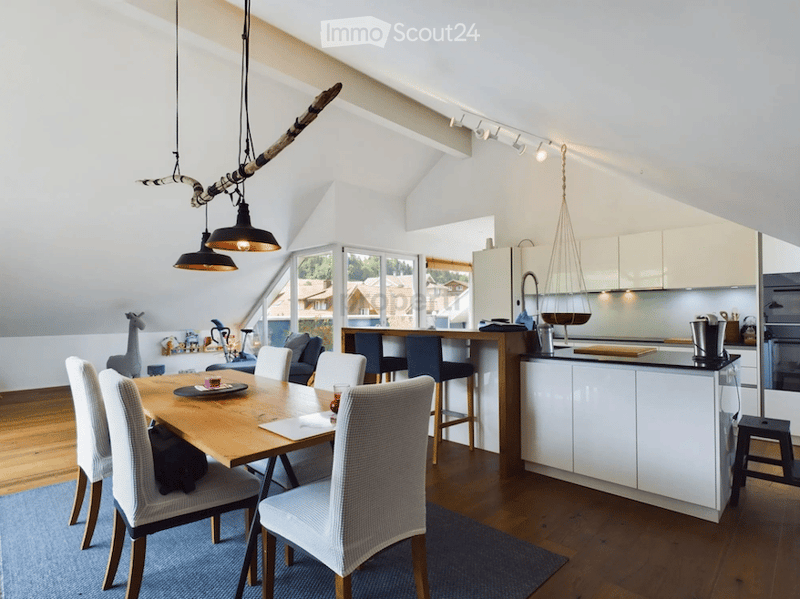 A modern, open-plan living and dining area with a wooden dining table, chairs, a kitchen island, and pendant lights. The space features white cabinets, a refrigerator, and a coffee machine, with a view of the outdoors through large windows.